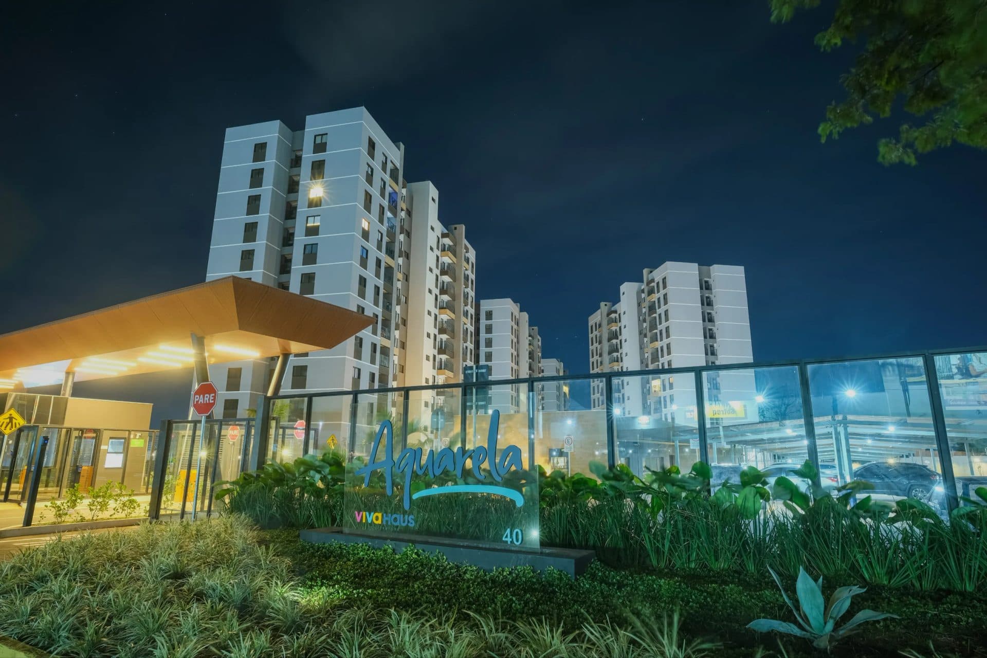 Fachada noturna iluminada da portaria e letreiro de identificação do condomínio Aquarela.
